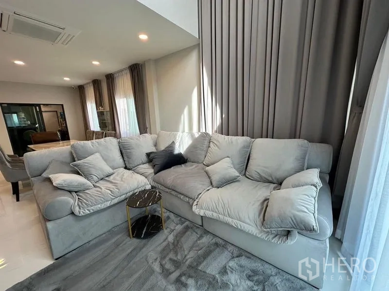 独立式住宅 出租在Bang Phli, Samut Prakan - 温馨起居角，柔软转角沙发、落地窗帘与灰色地毯上的小边几。