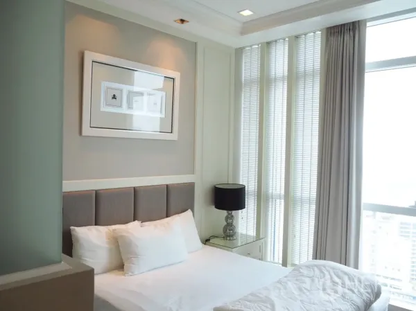 Condominium for rent in Pathum Wan, Bangkok - Cozy bedroom with upholstered headboard, bedside lamp, wall art and tall windows with blinds and curtains.