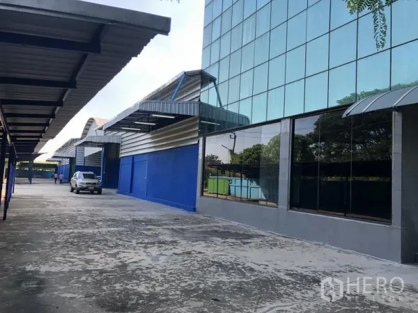 Warehouse for rent in Sai Mai, Bangkok - Modern glass-fronted office building beside blue steel warehouses and a wide driveway in Sai Mai.