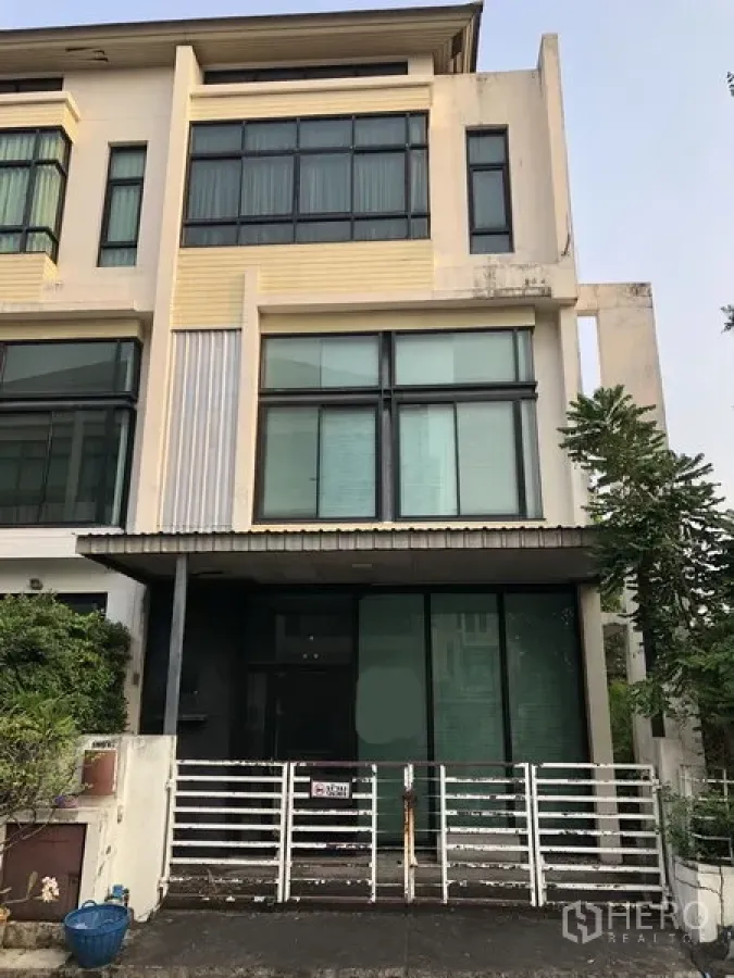Home Office for sale in Wang Thonglang, Bangkok - Modern multi-storey home office facade with tall glass windows, sliding doors and a gated entrance, with greenery at the side in Wang Thonglang, Bangkok.