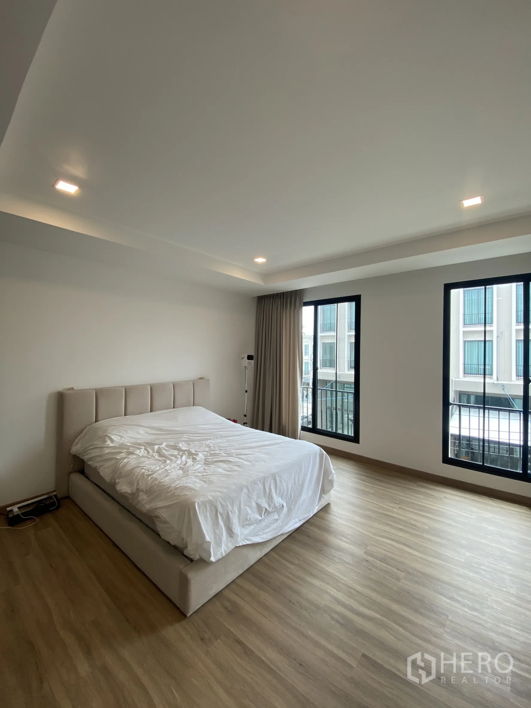Townhouse for rent or sale in Bang Khae, Bangkok - Large bedroom with upholstered bed, wood floor, and two tall windows