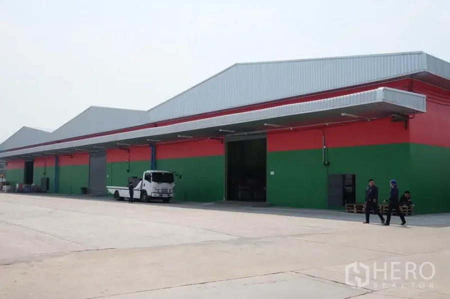 货栈 出租在Ban Phaeo, Samut Sakhon - 现代仓库外观，红绿立面与长金属屋顶，超大混凝土场地与多处装卸月台。