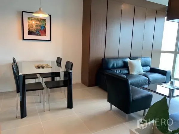 Condominium for rent in Khlong San, Bangkok - Open-plan living and dining area with a glass table, black leather sofa, wood wall panels and a large window.