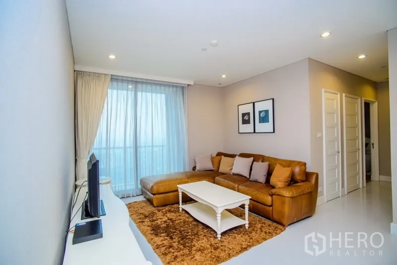 คอนโด ให้เช่าในKhlong Toei, Bangkok - ห้องนั่งเล่นสว่าง โซฟาหนังสีน้ำตาล โต๊ะกลางสีขาว ชุดทีวี และหน้าต่างเต็มผนังเปิดออกสู่ระเบียง