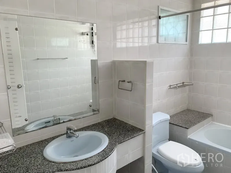 Detached House for rent in Khlong Toei, Bangkok - White-tiled bathroom with wide mirror, granite counter, toilet and bathtub with glass-block windows.