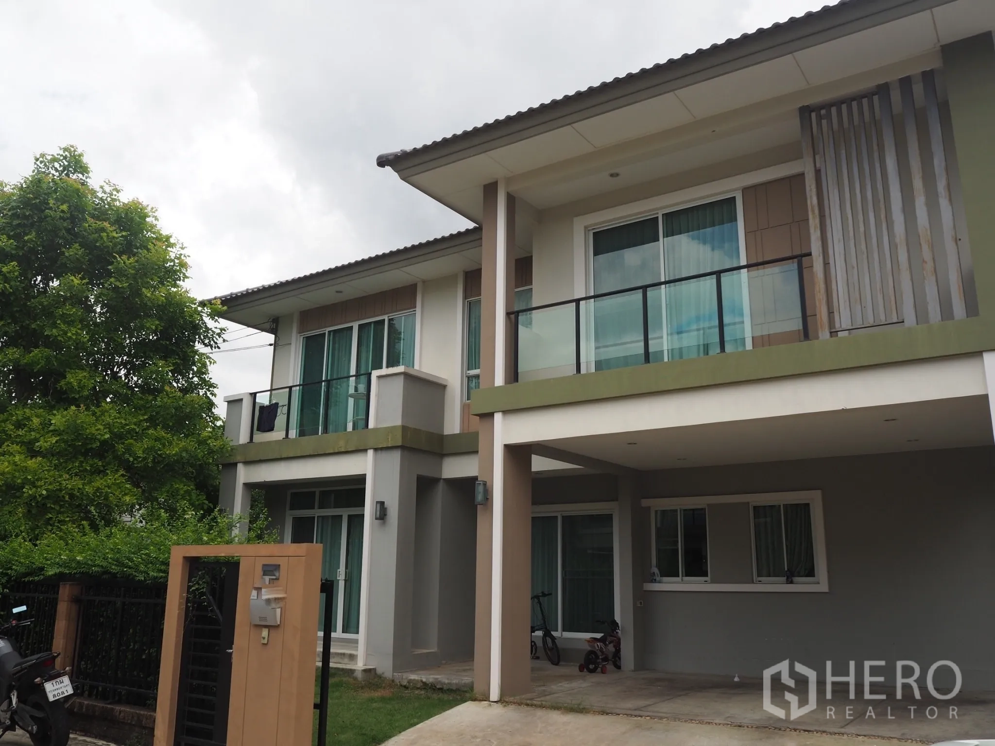 Detached House for rent in Pathum Thani - Two-storey detached house with balcony, covered carport, gated entry and mature trees.