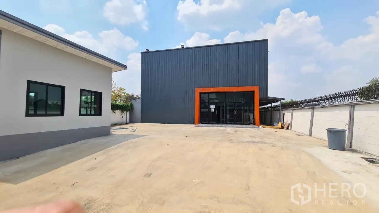 Home Office for rent in Pathum Thani - Modern home office façade with dark metal cladding, orange-framed glass entry and a wide concrete parking yard.
