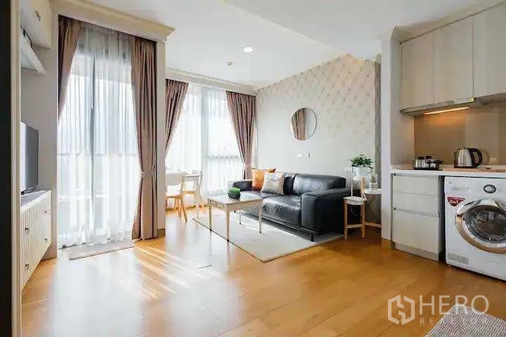 Condominium for rent in Khlong Toei, Bangkok - Bright open living area with wood floors, sofa, round wall mirror and kitchenette.