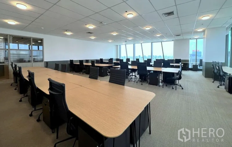 Office Space for rent - High-floor open-plan office with long workstations, ergonomic chairs, carpeted flooring, suspended ceiling lights, and floor-to-ceiling windows with Bangkok skyline views.