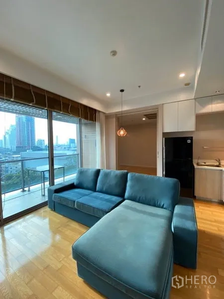 Condominium for rent or sale in Yan Nawa, Bangkok - Living room opening to a balcony with skyline view, blue sectional seating and adjacent kitchen.