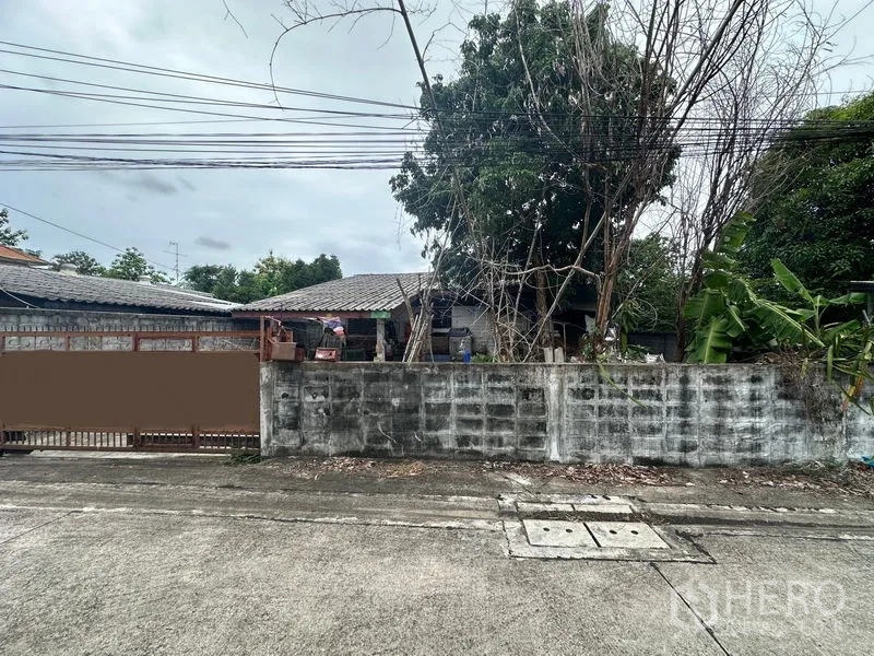 ที่ดิน ขายในPhra Khanong, Bangkok - ภาพมุมกว้างหน้าที่ดิน กำแพงเตี้ย บ้านอยู่หลังต้นไม้และสายไฟเหนือถนน