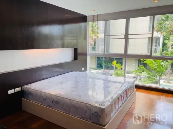 Condominium for rent in Watthana, Bangkok - Bright bedroom with platform bed, feature wall, and large windows facing greenery.