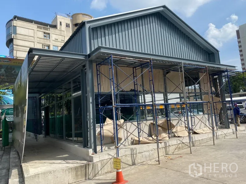 零售空间 出租在Khlong Toei, Bangkok - 商铺侧视图，带雨棚与落地窗，脚手架搭建中，前方宽阔出入口便于顾客与物流进出。