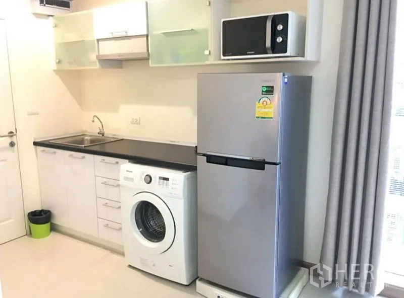 Condominium for rent in Phra Khanong, Bangkok - Compact kitchen with white cabinetry, black countertop, sink, in-unit washing machine, tall refrigerator and microwave by the window.