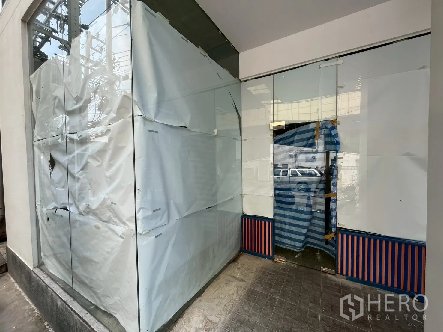 Retail Space for rent in Watthana, Bangkok - Corner retail unit with floor-to-ceiling wraparound glass and a taped glass door facing a paved walkway, with urban power lines visible outside.