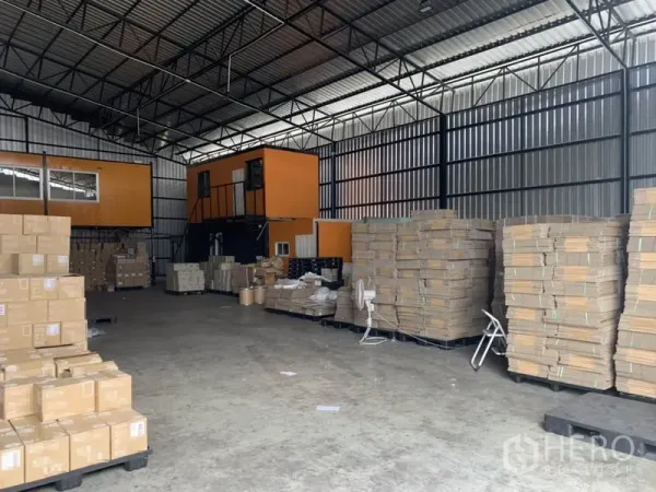 Warehouse for rent in Bang Kapi, Bangkok - Open-plan warehouse interior with a steel truss roof, concrete floor, and stacked pallets of cartons.