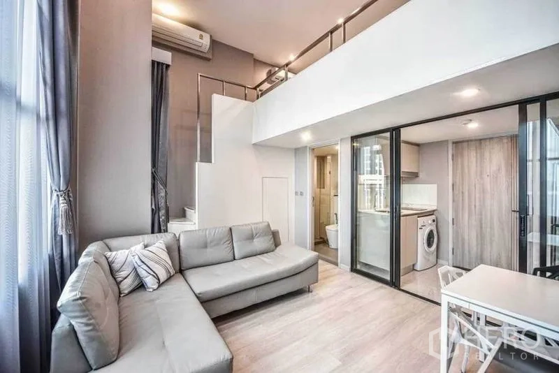 Condominium for rent in Sathon, Bangkok - Alternate angle of living room showcasing staircase to loft, sectional sofa and glass-partitioned kitchen.