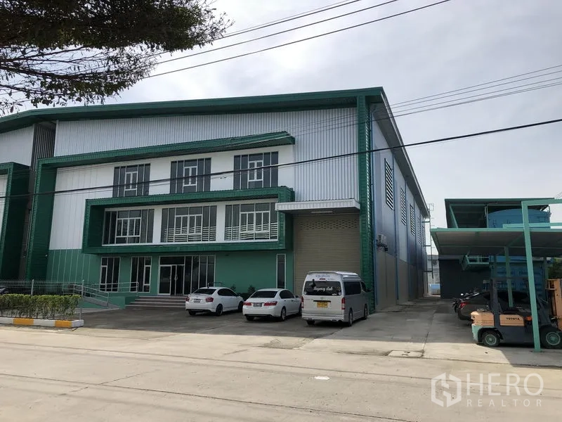 Factory for rent or sale in Bang Phli, Samut Prakan - Angled exterior view of the factory showing a wide driveway, loading door, and parked vehicles.