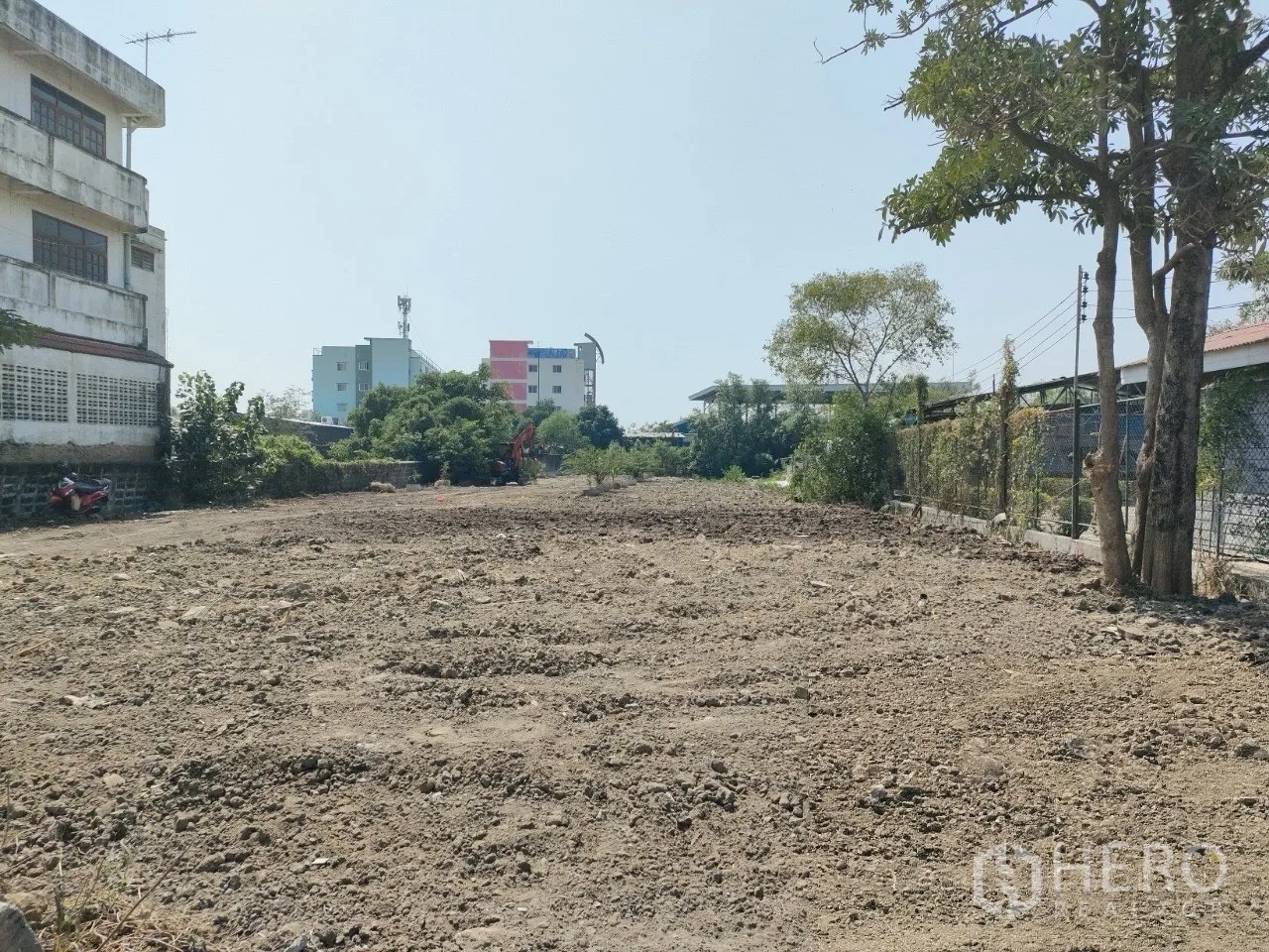 Land for sale in Mueang Samut Prakan, Samut Prakan - Level site with excavator in the distance and surrounding buildings beyond the trees.