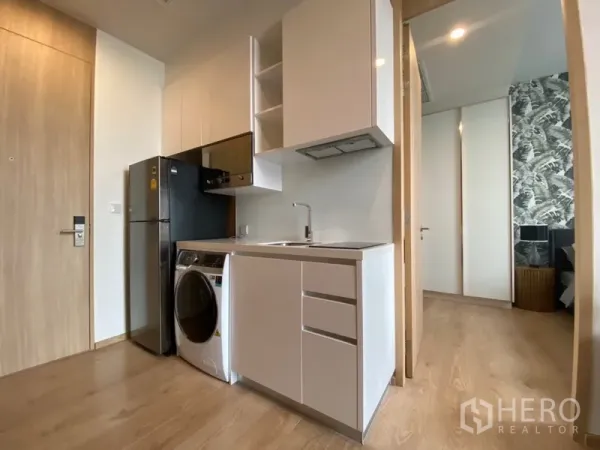 Condominium for rent in Watthana, Bangkok - Compact white kitchen with sink, hood, cooktop, fridge and in-unit washer beside the bedroom entrance.