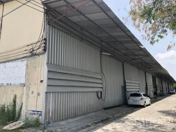 Warehouse for rent in Lam Luk Ka, Pathum Thani - Exterior row of metal warehouses with large sliding doors, covered loading bay and driveway with parked cars.