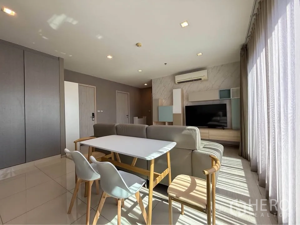 Condominium for rent in Khlong Toei, Bangkok - Living and dining space with sofa, table for four and media wall by tall windows.
