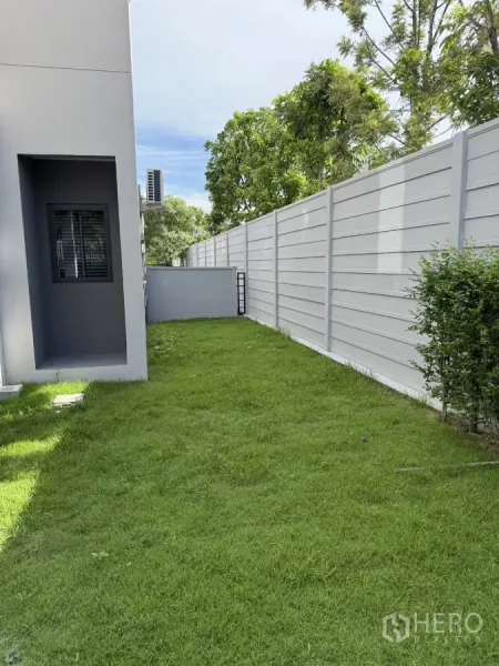 Townhouse for rent or sale in Bang Phli, Samut Prakan - Grassy backyard strip beside a tall grey boundary fence and house wall.