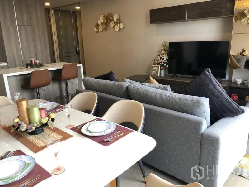 Condominium for rent in Watthana, Bangkok - Open-plan living and dining area with a gray sofa, TV wall, and a set dining table beside the kitchen.