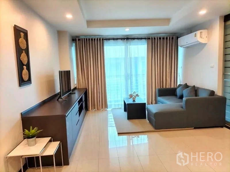 Condominium for rent in Watthana, Bangkok - Bright living room with L-shaped gray sofa, TV console, air conditioner and balcony with floor-to-ceiling curtains.