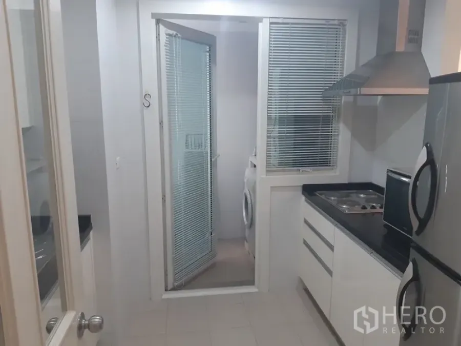 คอนโด ให้เช่าในKhlong Toei, Bangkok - ครัวปิดสว่างพร้อมท็อปสีดำ เตาและเครื่องดูดควัน ประตูกระจกเปิดสู่โซนซักล้างที่มีเครื่องซักผ้า