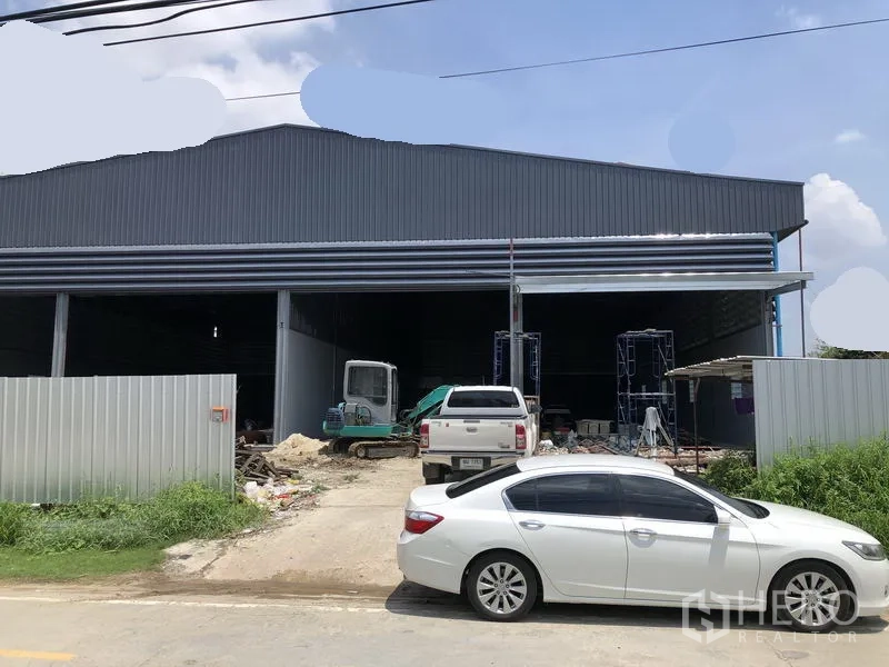 Warehouse for rent in Bang Khen, Nonthaburi - New steel-clad warehouse facade with wide roller doors, driveway and parked cars.
