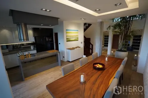 Townhouse for rent in Khlong Toei, Bangkok - Open-plan dining with a long wood table beside a modern island kitchen, indoor greenery and staircase.