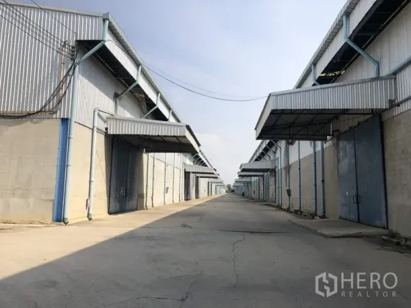 货栈 出租在Saraburi - 成排工业仓库外立面，宽阔车道配金属大门与雨棚，便于卡车通行。