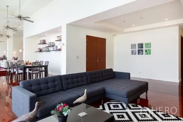 Condominium for rent in Bang Rak, Bangkok - Open-plan living room with dark sectional sofa, patterned rug, polished wood floors and dining area in the background.