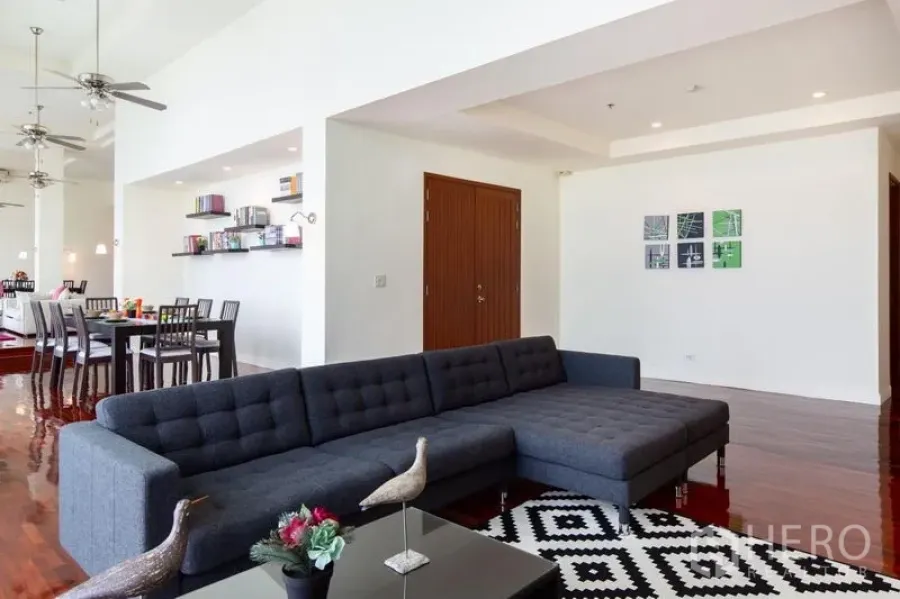 Condominium for rent in Bang Rak, Bangkok - Open-plan living room with dark sectional sofa, patterned rug, polished wood floors and dining area in the background.