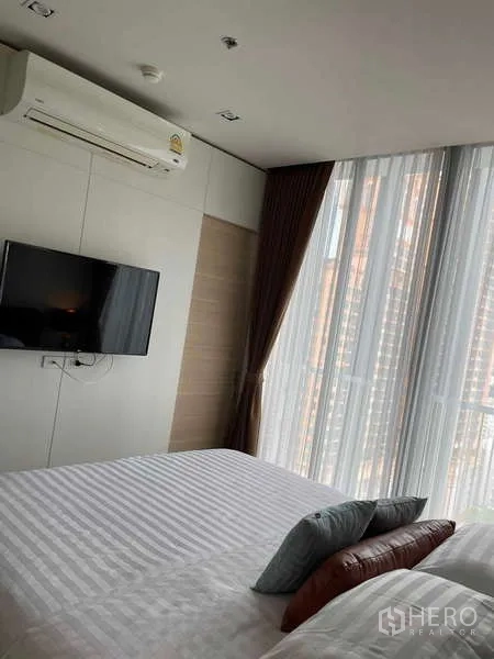 Condominium for rent or sale - Bedroom corner with wall-mounted TV, air conditioner and tall sheer curtains over a city-view window.
