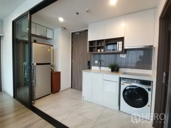 Condominium for rent in Huai Khwang, Bangkok - Compact white kitchen with built‑in cabinets, sink, microwave, fridge and front‑loading washer.