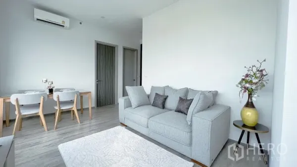 Condominium for rent in Watthana, Bangkok - Minimalist living room with gray sofa, air conditioning, dining nook, and doors leading to two bedrooms.