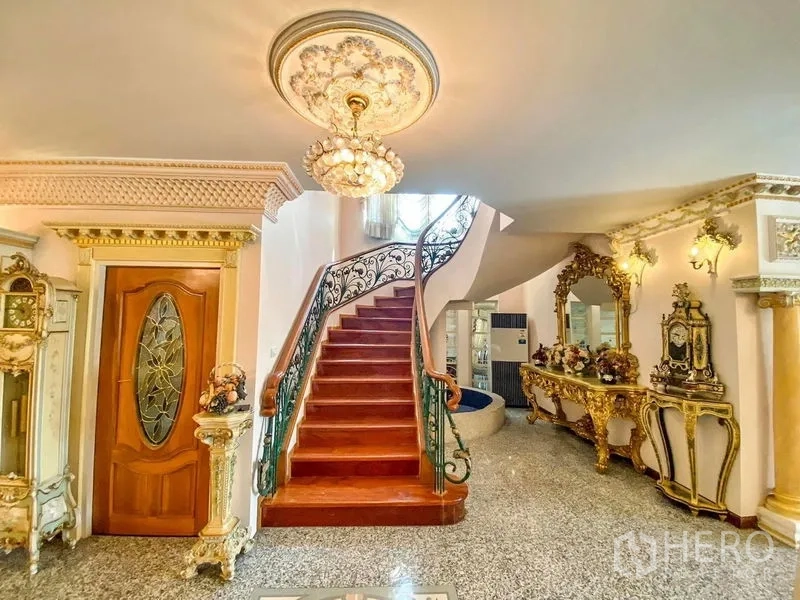 独立式住宅 出租或出售在Bang Khen, Bangkok - 弧形木楼梯搭配锻铁栏杆，金色玄关桌与吊灯点缀其间。