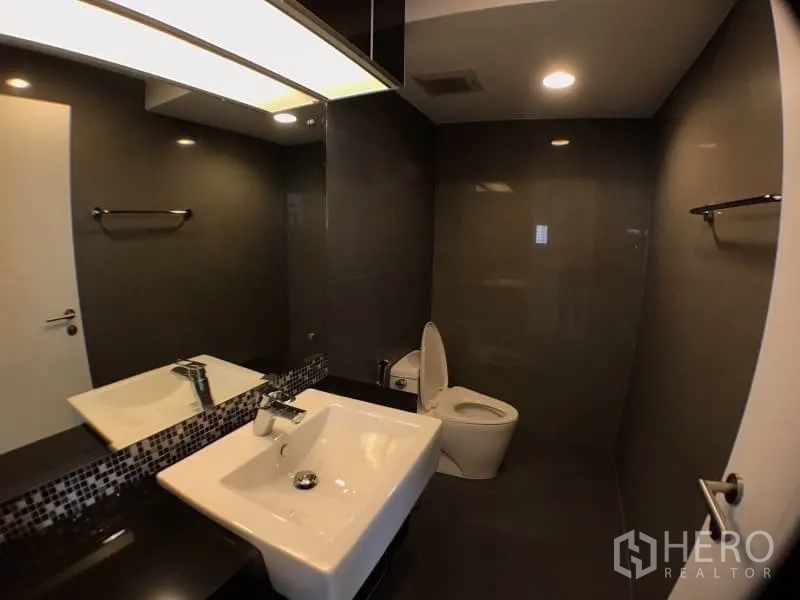 Condominium for rent in Khlong Toei, Bangkok - Modern bathroom with dark tiles, large mirror, vanity and toilet.