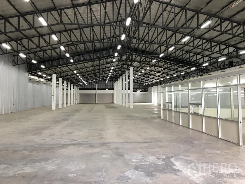 货栈 出租在Nonthaburi - 宽阔仓库地面，左侧金属包墙，右侧为办公室区块。