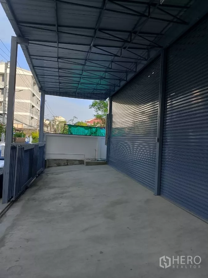 Warehouse for rent in Huai Khwang, Bangkok - Side driveway and roofed loading area beside grey roller shutters.