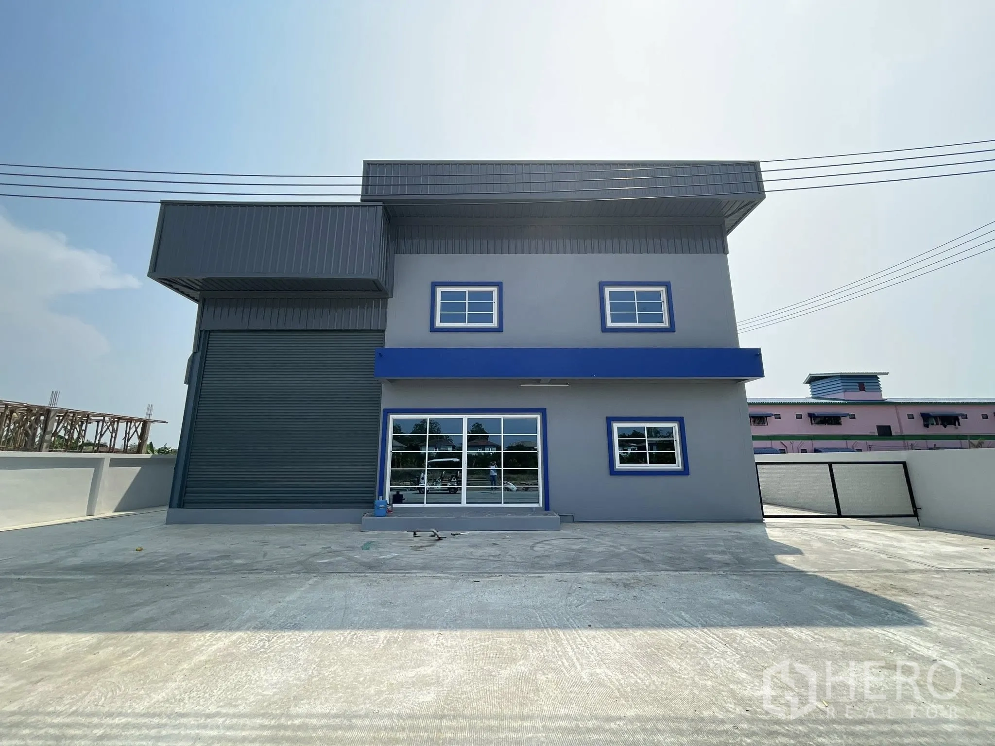 Factory for rent or sale in Bang Phli, Samut Prakan - Front exterior of a modern two-storey factory with large roller shutter, blue awning, and spacious concrete yard.