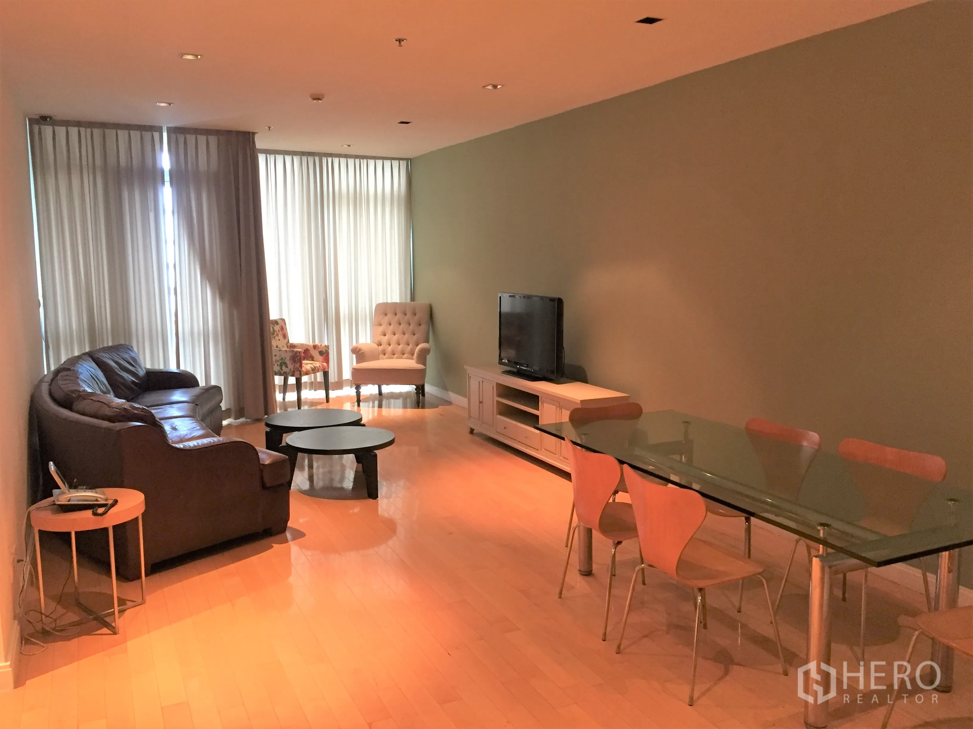 Condominium for rent or sale in Pathum Wan, Bangkok - Spacious living and dining room with leather sofa, glass table, TV console and floor-to-ceiling curtains on light wood flooring.