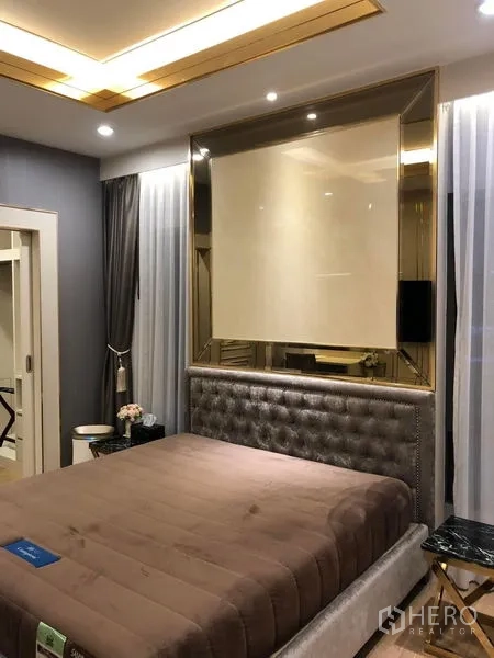 Detached House for rent or sale in Prawet, Bangkok - Contemporary bedroom with tufted headboard, mirrored accent wall, and soft lighting.