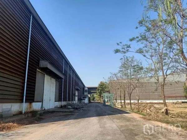 Warehouse for rent in Mueang Samut Sakhon, Samut Sakhon - Side lane beside corrugated warehouse walls with loading doors and clear blue sky.