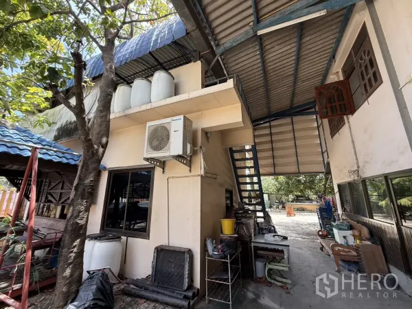 Warehouse for sale in Phra Samut Chedi, Samut Prakan - Side of house within the compound featuring external stair, AC unit and covered work area.
