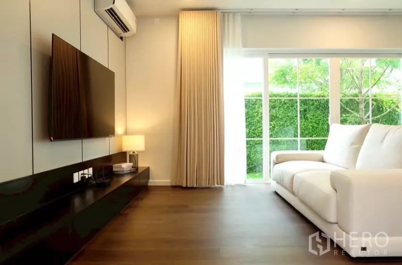 Detached House for rent in Bang Phli, Samut Prakan - Cozy living room with white sofa, media console and TV, sliding glass doors opening to a green garden.
