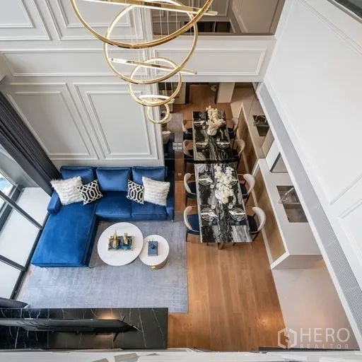 คอนโด ให้เช่าในKhlong Toei, Bangkok - มุมมองจากด้านบนของโถงนั่งเล่นเพดานสูงพร้อมโซฟาสีน้ำเงิน โต๊ะทานหินอ่อน พื้นไม้ และโคมไฟทรงวงกลมสีทอง