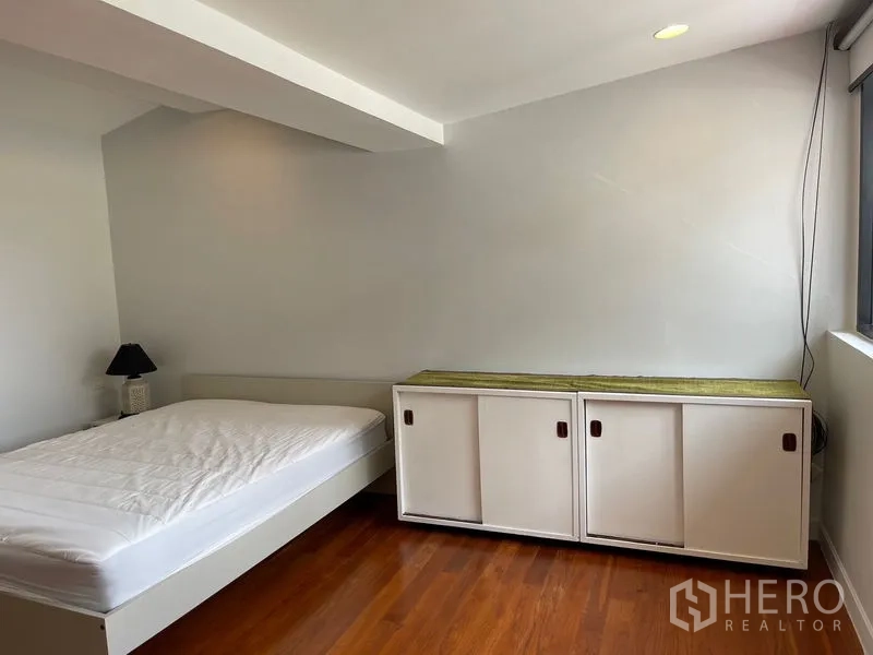 Townhouse for rent in Khlong Toei, Bangkok - Simple bedroom with wood flooring, low bed and built-in storage beside a window.